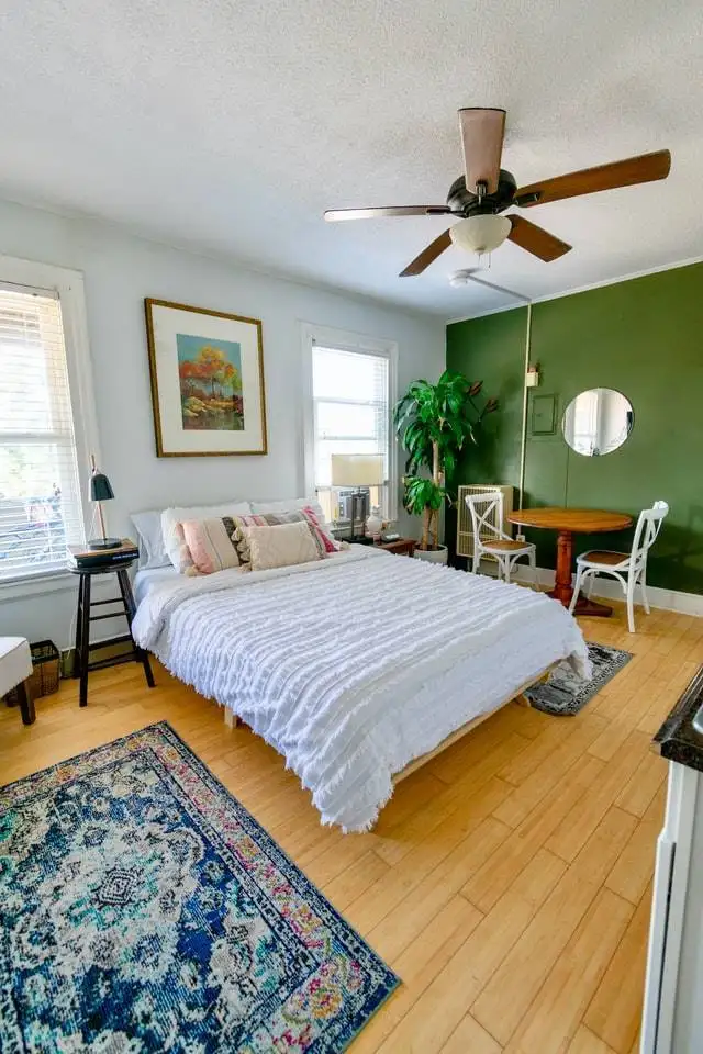 A cozy and well-lit bedroom with a mix of vintage and modern decor, featuring a prominent green accent wall, hardwood floors, and a ceiling fan.