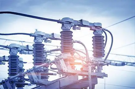 Electrical insulators and connections at a power substation with overhead power lines against a blue sky.
